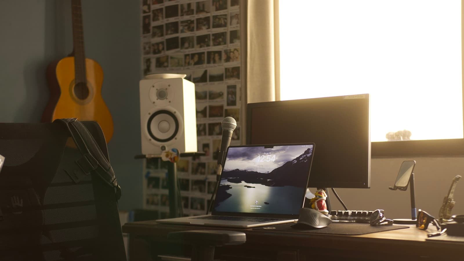 Home studio desk with a laptop, studio monitor, microphone, and an acoustic guitar on the wall